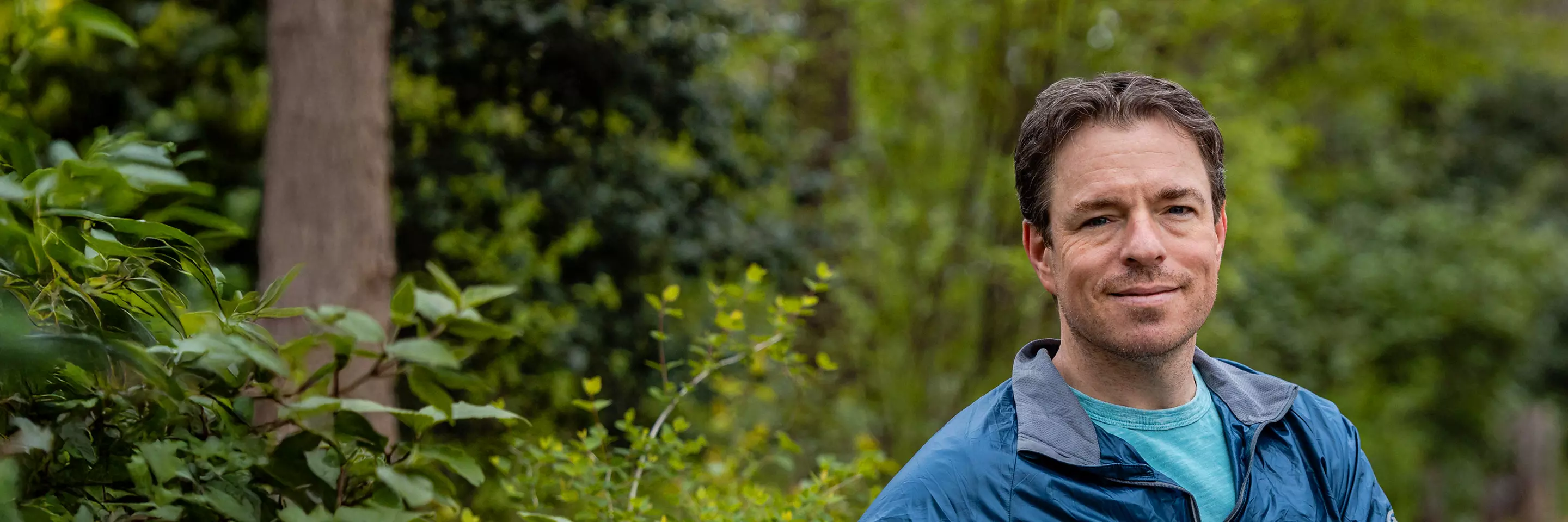 MSK patient Kieran, wearing a blue jacket, sits on a wooden bench surrounded by greenery, looking at the camera with a calm expression.
