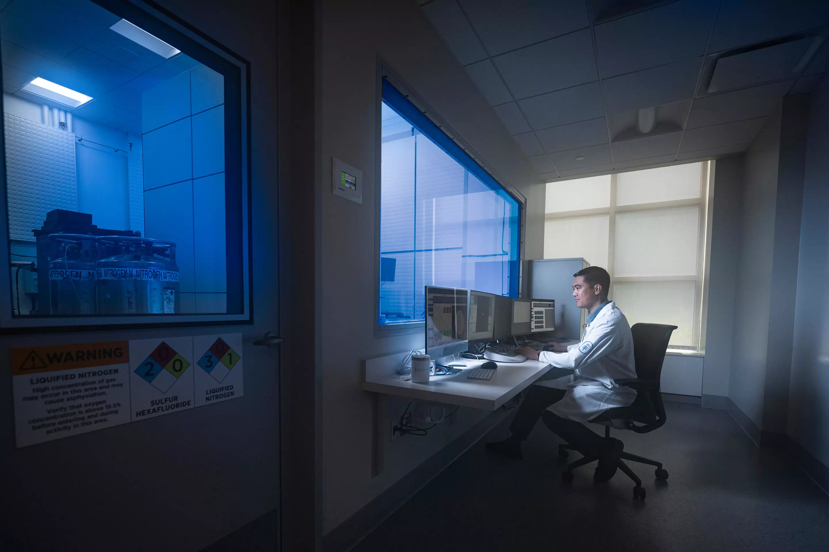 A person in a lab coat works at a computer desk, observing equipment through a window in a research facility.